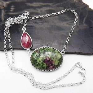 May include: A silver chain necklace with a green and pink oval pendant and a teardrop-shaped pendant. The oval pendant has a silver bezel setting and the teardrop pendant has a faceted design.