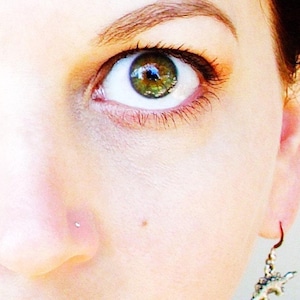 May include: Close-up of a person's face with green eyes and a small silver stud in their nose. The person is wearing a silver earring with a dinosaur charm.