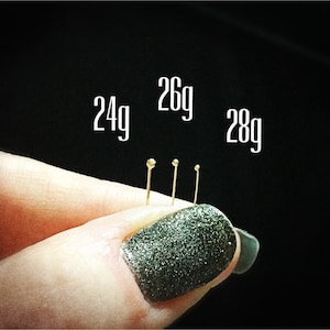 May include: Four gold-colored needles of different sizes, labeled 24g, 26g, and 28g. The needles are held in a hand with a dark green nail polish.