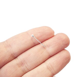 May include: A silver pin with a round head. The pin is held between two fingers.