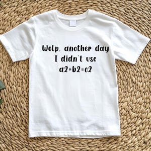 Puede incluir: Una camiseta blanca con el texto "Welp, another day I didn't use a2+b2=c2" en negro. La camiseta se muestra sobre una superficie tejida de color marrón claro con hojas verdes en la esquina.