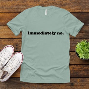 以下が含まれることがあります： 「Immediately no.」の文字が黒でプリントされた、薄い緑色のTシャツ。赤いアクセントの白いスニーカーと、木製の表面に置かれた小さな鉢植えの植物が添えられています。