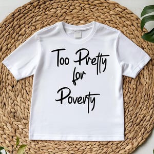 May include: White t-shirt with the phrase "Too Pretty for Poverty" in black script. The shirt is displayed on a woven, circular mat with green foliage in the background. The t-shirt has short sleeves and a crew neck.
