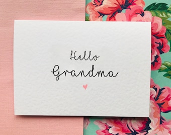 Puede incluir: Una tarjeta blanca con el texto "Hello Grandma" en escritura cursiva negra y un pequeño corazón rosa.