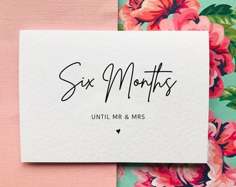 May include: A white card with black text that reads "Six Months Until Mr & Mrs" with a small heart symbol below the text. The card is on a pink and floral background.