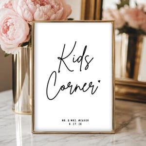 May include: A vertical rectangular sign in a thin gold frame featuring the black handwritten text Kids Corner with a small heart symbol, followed by the printed text MR. & MRS. WEAVER and the date 6. 27. 26 on a plain white background.
