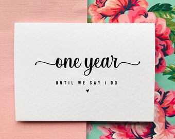 May include: White card with black text that reads "one year until we say I do" with a small heart symbol.