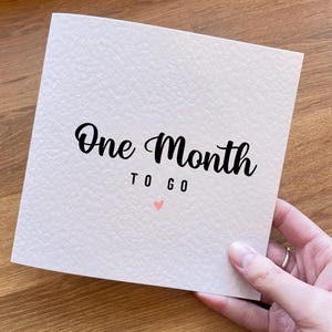 May include: A square, off-white greeting card with a textured surface. The card features the black text "One Month" in a script font, followed by "TO GO" in a sans-serif font, and a small, light pink heart icon centered below the text.