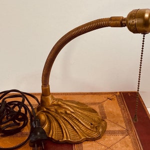 Puede incluir: Una lámpara de escritorio dorada con un brazo flexible y una base decorativa con forma de concha. La lámpara tiene un cable y un enchufe negros.