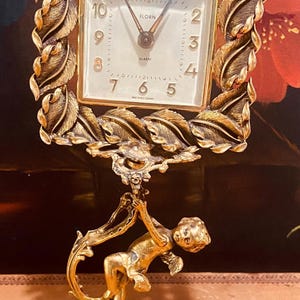 May include: A vintage gold-toned alarm clock with a square face and leaf-like frame. The clock face is white with black numerals and hands. The clock is supported by a decorative stand featuring a small figure. The clock reads "FLORN".