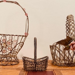 Puede incluir: Tres cestas decorativas de alambre. La más grande tiene un asa con cuentas rosas y un diseño floral. La más pequeña es una cesta en miniatura. La tercera cesta tiene un asa con un diseño tejido y una rosa rosa.