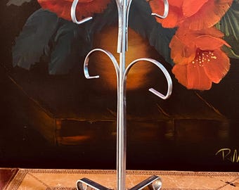 May include: A silver-colored metal mug tree with a four-legged base and curved arms for hanging mugs. The mug tree is displayed in front of a painting of large red flowers.