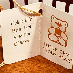 May include: A white tag with gold lettering that reads "Collectible Bear Not Safe For Children" and "Little Gem Teddy Bear". The tag is attached to a wooden base with a small, brown teddy bear illustration.
