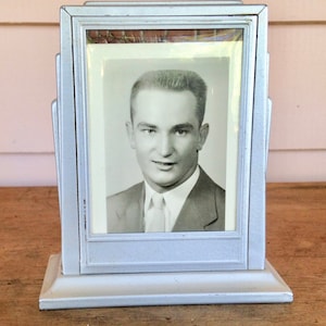 Puede incluir: Un marco de fotos plateado con una foto en blanco y negro de un hombre en su interior. El marco tiene un diseño decorativo y una base.