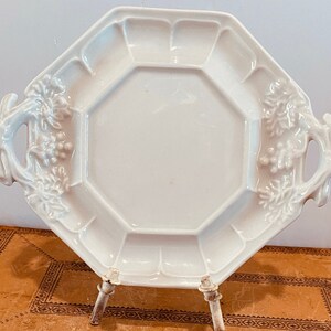 Puede incluir: Bandeja de servir de porcelana blanca con forma octogonal y dos asas. Las asas están esculpidas para parecerse a ramas con hojas y uvas.