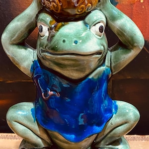 May include: A green ceramic frog figurine wearing a blue shirt, holding a brown ceramic pot above its head. The frog is sitting on a brown base.