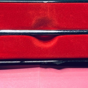 May include: A silver-colored instrument with a T-shaped design, resting in a red velvet-lined case. The instrument has a central bar with a knob and a perpendicular arm. The case is black with a pink paper underneath.