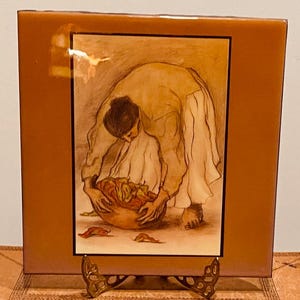 Puede incluir: Un azulejo de cerámica con un borde marrón y una imagen enmarcada de una mujer con un vestido blanco sosteniendo una cesta de fruta. La mujer se inclina sobre la cesta y su rostro está oculto por su cabello. La imagen está pintada en tonos marrones y beige.