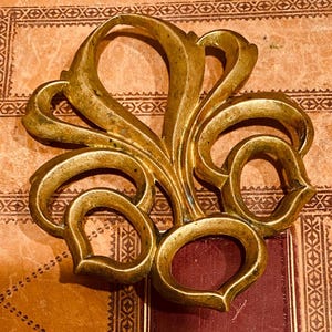 May include: A gold-colored metal decorative piece with a stylized floral design. The piece has a smooth, polished finish and intricate details.