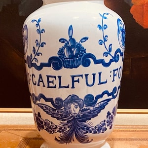 Peut inclure: Un vase en céramique blanc avec des éléments décoratifs bleus. Le vase présente un motif floral bleu, un chérubin ailé et le texte "CAELFUL FO". Le vase a une base large et un bord légèrement évasé.