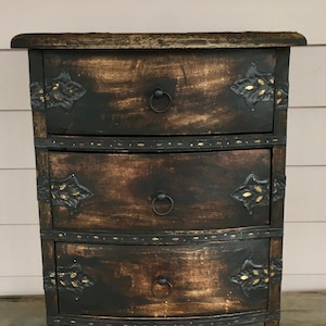 Puede incluir: Un arcón de madera marrón oscuro con tres cajones. Cada cajón tiene una manija de metal y detalles decorativos de metal. El arcón tiene un acabado desgastado.