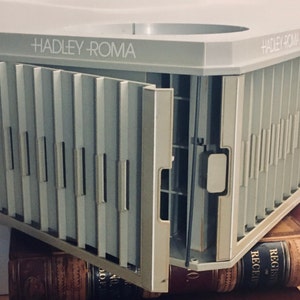 Könnte beinhalten: Ein grauer Plastik-Kartenhalter mit dem Text "HADLEY ROMA" auf der Oberseite. Der Halter hat mehrere Fächer für Karten und ist geöffnet, um die Fächer zu zeigen.