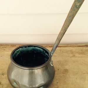 May include: A vintage copper pot with a long handle. The pot has a rounded bottom and a slightly flared rim. The handle is made of metal and is attached to the pot with a rivet. The pot is tarnished and has a green patina.