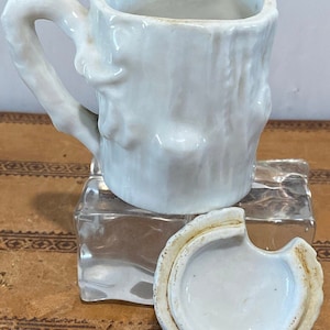 May include: White ceramic mug shaped like a tree trunk with a handle that looks like a branch. The mug has a lid that is also shaped like a tree trunk. The mug is sitting on a clear glass block.