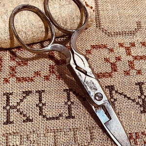 Può includere: Forbici da cucito Singer vintage con finitura argentata. Le forbici hanno grandi fori per le dita e una lama appuntita. Il testo "SINGER" è visibile sulle forbici. Sono appoggiate su una superficie beige strutturata.