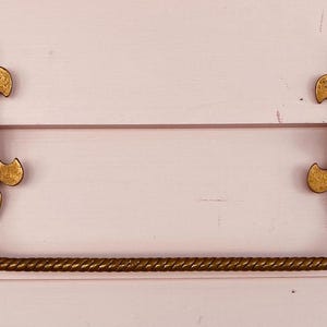 Op de afbeelding: Een gouden, decoratieve handdoekhouder met een gedraaid touwontwerp en sierlijke fleur-de-lis-eindstukken. De stang is gemonteerd op een lichtroze muur.