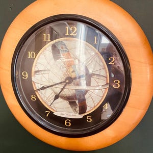 May include: Round wall clock with a light wood frame and a black inner ring. The clock face features a world map design with gold numerals and hands. The text "Atlas Coelestis - 1660" is visible.