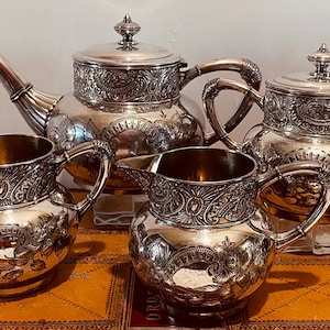 May include: A silver tea set with detailed floral and scrollwork. The set comprises a teapot, sugar bowl, creamer, and two handled cups. The items are displayed on a brown surface.