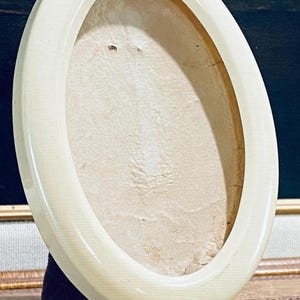 May include: An off-white, oval-shaped picture frame with a glossy finish. The frame stands on two small, rounded feet. The interior of the frame holds a light-colored backing, suggesting it is designed to hold a photograph or artwork.