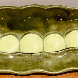 May include: A green ceramic pea pod serving dish with six individual compartments. The dish is glazed in a glossy green finish and has a realistic pea pod design.