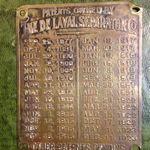Peut inclure: Une plaque en laiton avec le texte "PATENTS OWNED BY THE DE LAVAL SEPARATOR CO." et une liste de dates et de numéros de brevets. La plaque est usée et a un fond vert.