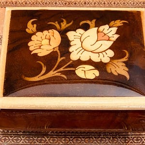 May include: A wooden music box with a floral inlay design. The box is brown with a light brown trim and a clear lid. The floral design features a white rose with a pink center and green leaves.