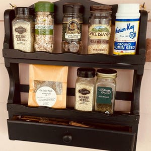 May include: A black wooden spice rack with two shelves and a small drawer. The top shelf holds jars of sesame seeds, crystallized ginger, allspice, and cumin seed. The bottom shelf has sesame seeds, dill weed, and a spice pack.