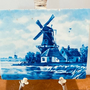 May include: Blue and white Delft tile depicting a windmill and houses in a Dutch landscape. The tile is on a white easel.