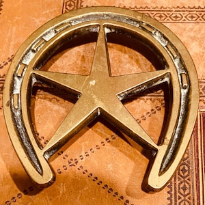 Op de afbeelding: Een messingkleurig hoefijzervormig object met een vijfpuntige ster in het midden. Het hoefijzer heeft een textuur en staat tegen een bruine achtergrond met decoratieve patronen. De ster is gecentreerd in het hoefijzer.