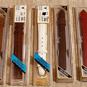 Puede incluir: Una colección de correas de reloj vintage en varios colores, incluyendo rojo, negro, marrón y blanco, exhibidas en su embalaje original. Las correas son de cuero y resistentes al agua.