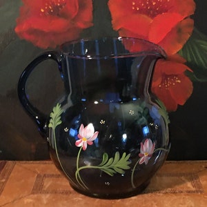 May include: A cobalt blue glass pitcher with a hand-painted design of white and pink flowers with green leaves. The pitcher has a round body and a handle.