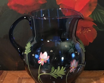 以下が含まれることがあります： 手描きの白とピンクの花と緑の葉のデザインが施されたコバルトブルーのガラスピッチャー。ピッチャーは丸いボディとハンドルが付いています。