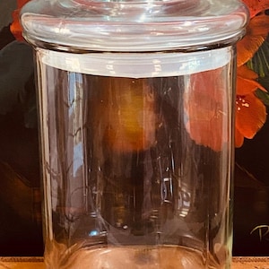 May include: A clear glass apothecary jar with a lid. The jar is cylindrical with a rounded base and a clear, round lid with a small knob. The background features a dark surface with red and orange floral accents.