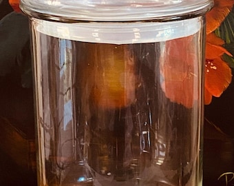 May include: A clear glass apothecary jar with a lid. The jar is cylindrical with a rounded base and a clear, round lid with a small knob. The background features a dark surface with red and orange floral accents.