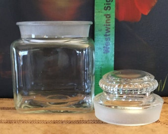 May include: A clear glass jar with a frosted lid. The jar is square shaped and has a wide mouth. The lid has a raised, decorative rim.