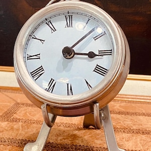 May include: A silver-coloured, round clock with a white face and black Roman numerals. The clock has a small loop at the top and stands on three curved legs. The hands are black, and the clock face is protected by a clear cover.