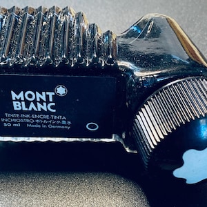 Peut inclure: Flacon d'encre Montblanc noir avec un dessus cannelé et un logo blanc sur le bouchon. L'étiquette indique "MONTBLANC" et "TINTE-INK-ENCRE-TINTA INCHIOSTRO ボトルインク・墨水 50 ml Made in Germany."