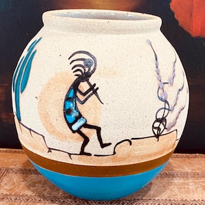 Puede incluir: Un jarrón de cerámica blanca con un diseño pintado en azul, marrón y negro de un bailarín nativo americano. El jarrón tiene un cuerpo redondo y una base azul.