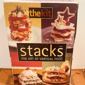 May include: A book titled "stacks: the art of vertical food" by "the kit". The cover features four photos of stacked food, including a salad, a dessert, a rice dish, and a layered pastry.
