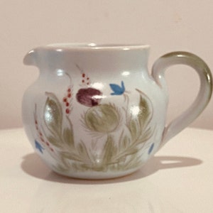 May include: A white ceramic creamer with a green handle and a floral design featuring a purple flower, green leaves, and blue accents.